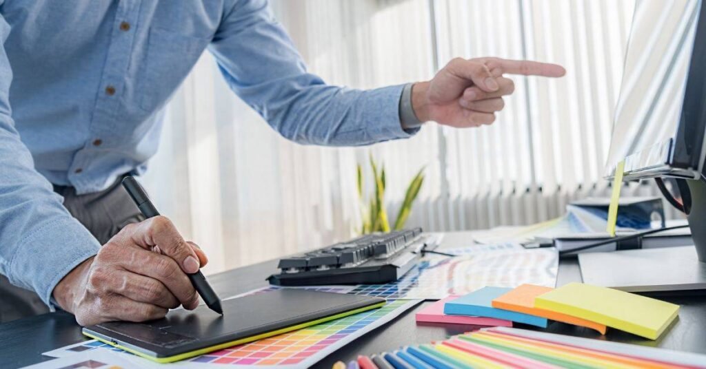 Designer using tablet and computer workspace with color samples and creative tools.