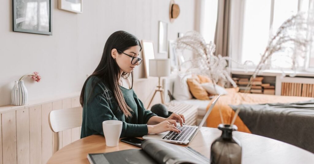 Young woman working on a laptop at a desk in a cozy home environment with books, coffee, and soft natural light.