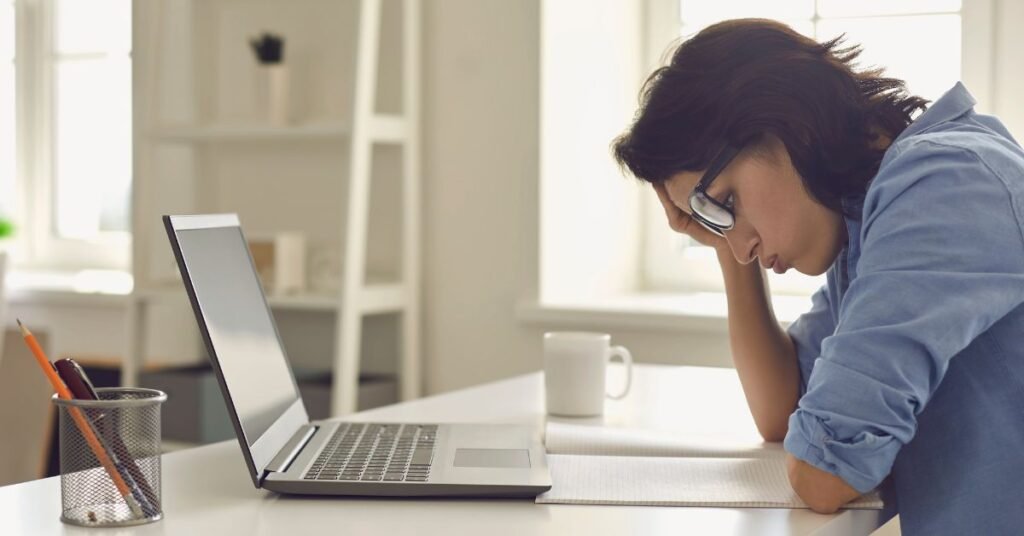 Woman feeling stressed while working on laptop at home office desk with notebook and coffee mug nearby